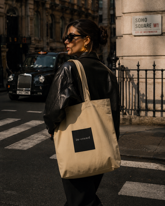 Person holding a beige tote bag with 'in-crowd' logo on a city street.