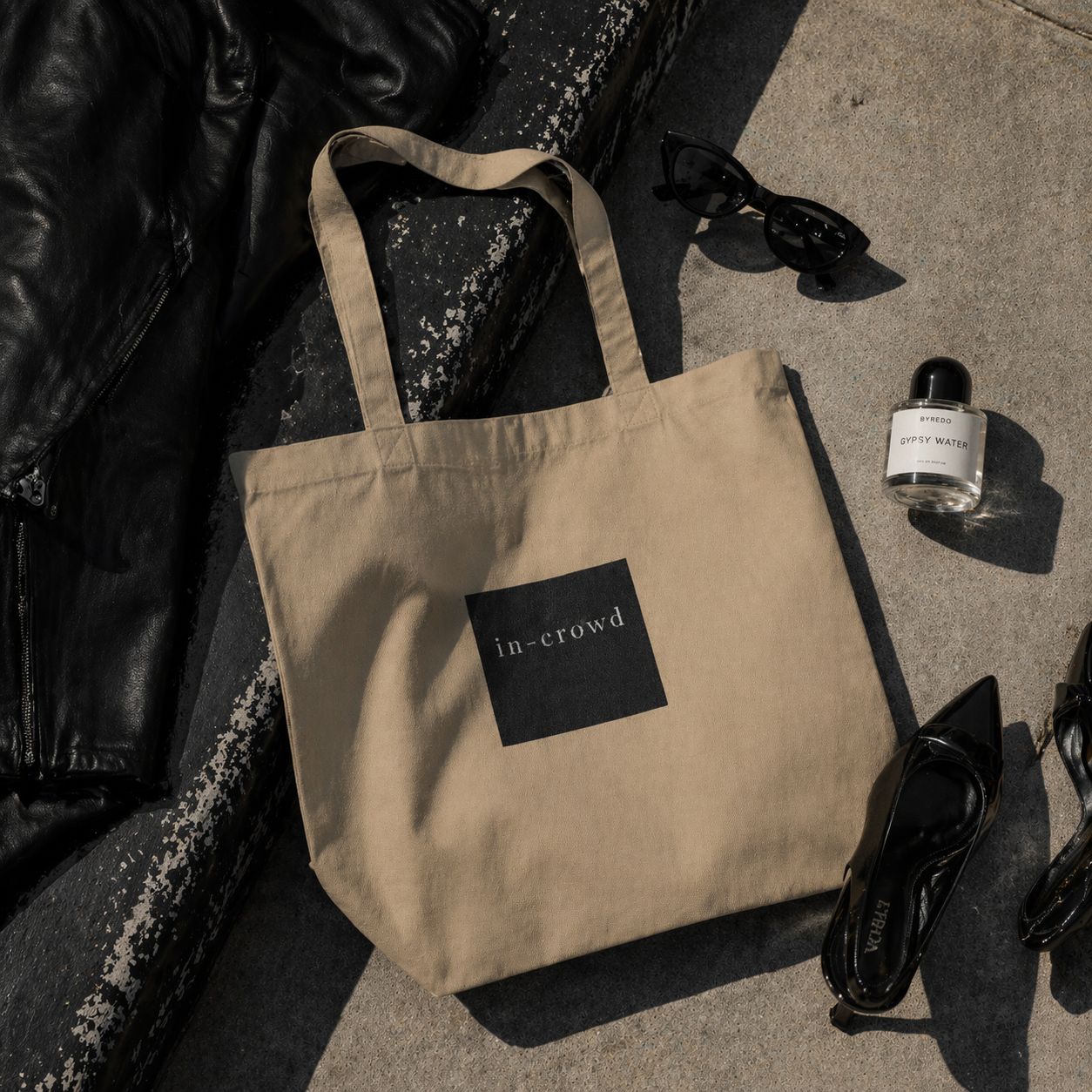 Beige tote bag with 'in-crowd' logo on a concrete surface with a black jacket and sunglasses.