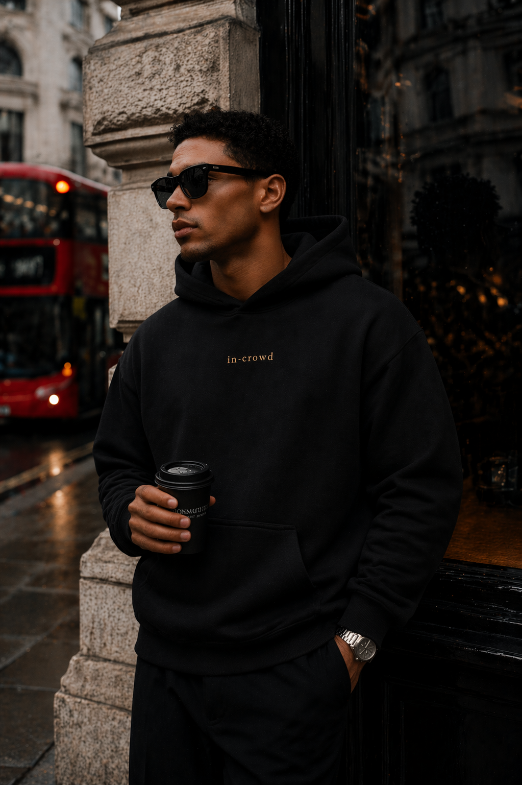 Man wearing a black hoodie with text, holding a coffee cup, standing in an urban setting.