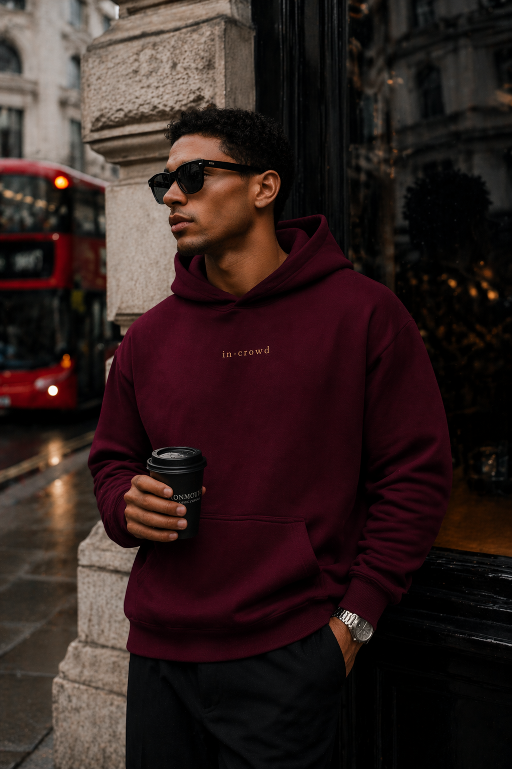 Man wearing a burgundy hoodie with 'in crowd' text, holding a coffee cup, standing in an urban setting.