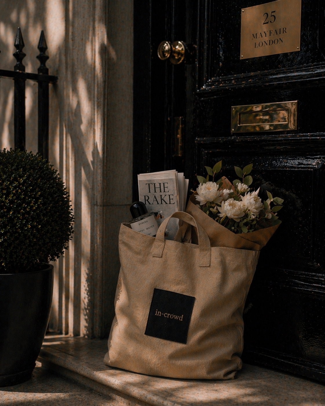 Beige tote bag with 'in-crowd' logo on a doormat in front of a black door with gold handle and plaque.