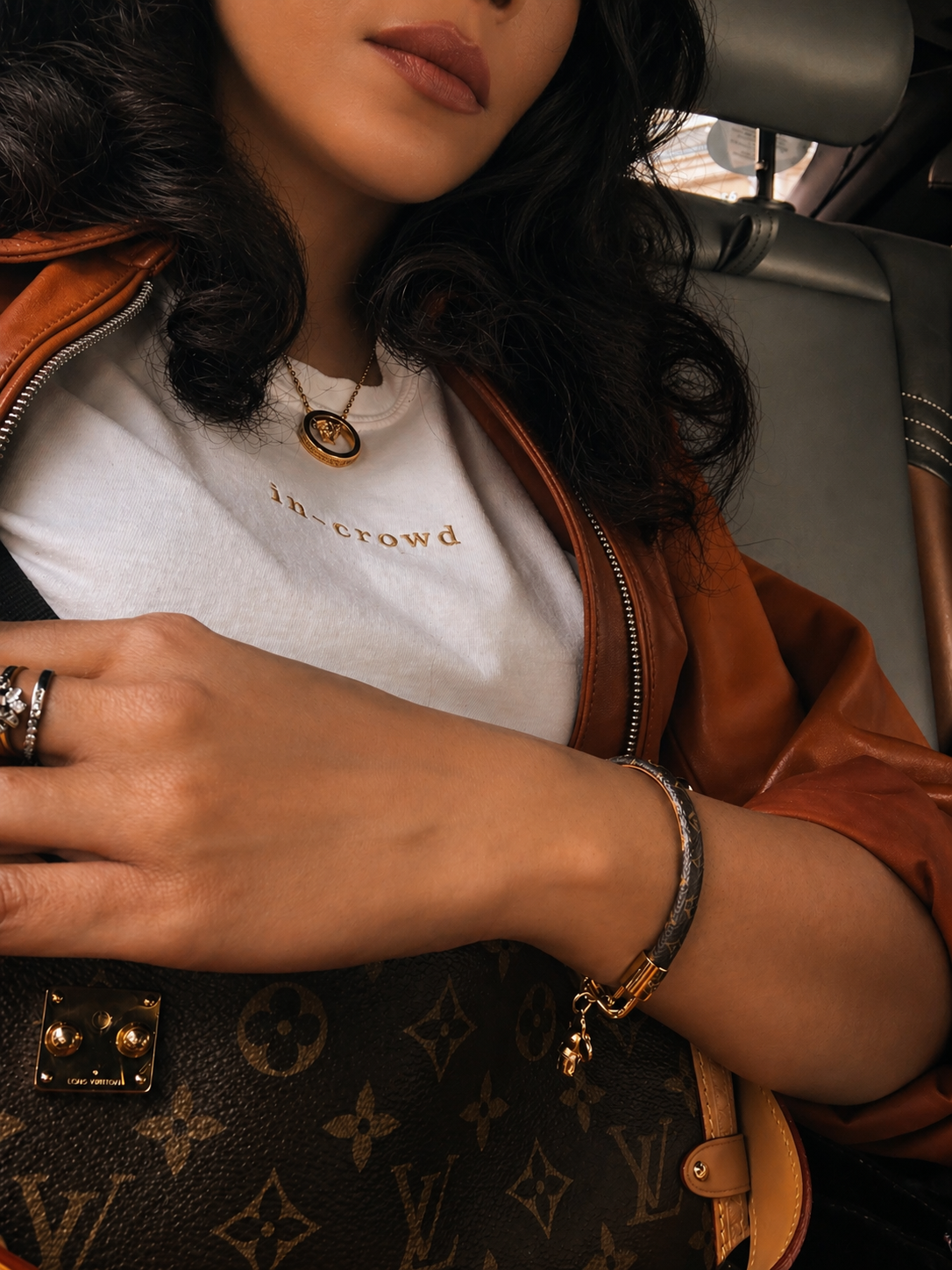 Person wearing a white shirt with text, brown jacket, and holding a Louis Vuitton handbag.