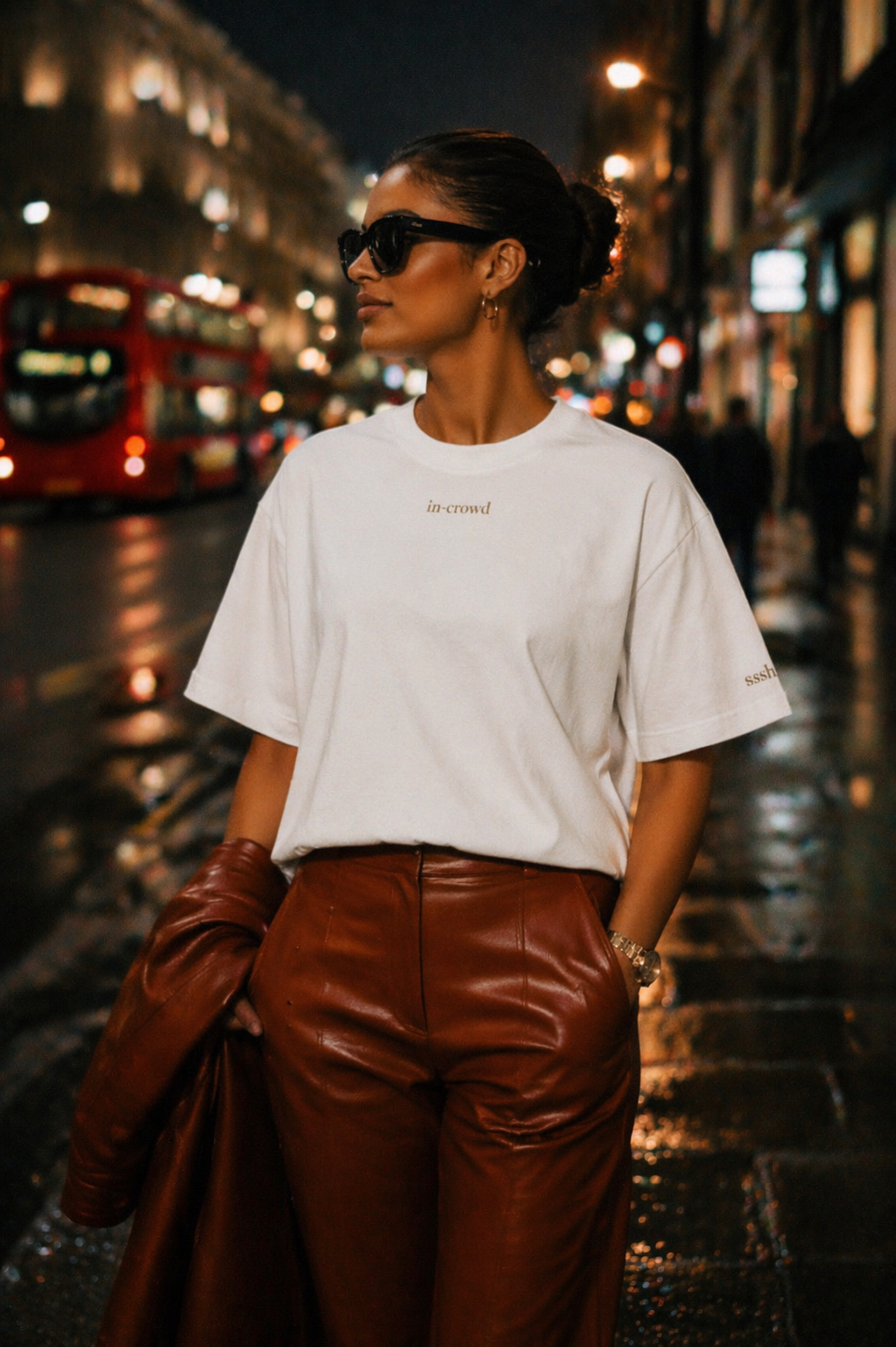 Woman in white t-shirt and brown leather pants standing on a city street at night.