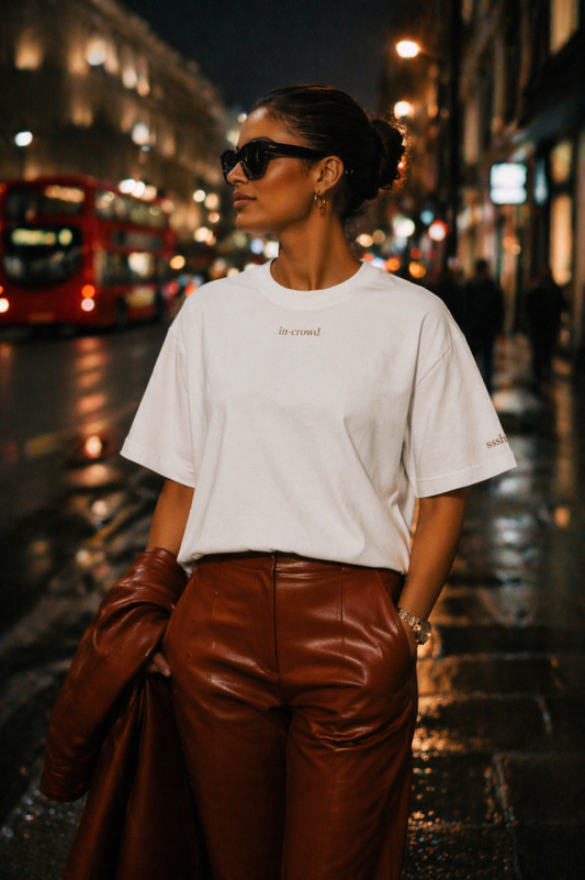 Woman in white t-shirt and brown leather pants standing on a city street at night.